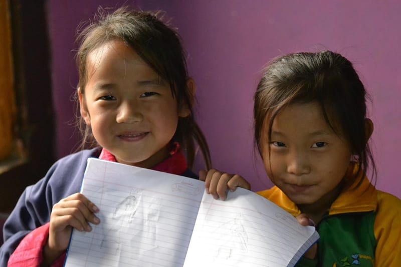 Two smiling schoolgirls holding up an open notebook with their pencil drawings